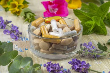 Çeşitli kapsüller ve tabletler canlı çiçekler ve yeşilliklerle çevrili cam bir kasede düzenlenerek, sağlık ve doğa arasındaki uyumlu bağlantıyı gösteriyor..