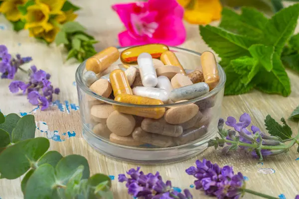 Çeşitli kapsüller ve tabletler canlı çiçekler ve yeşilliklerle çevrili cam bir kasede düzenlenerek, sağlık ve doğa arasındaki uyumlu bağlantıyı gösteriyor..