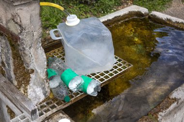 Temiz bir kap, bireyler onu suyla doldurduğu için doğal bir su kaynağının yanında durur. Yakınlarda, yeşil bantlı şişeler kolay taşınma için hazırlanıyor. Mekan huzurlu ve manzaralı..