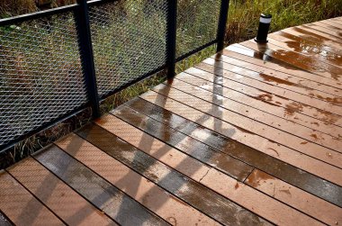 Yağmur sonrası parktaki ıslak ahşap zemin yolu yatay oranda izole edilmiş demir metal ağlar akşam güneşinin arka planına sahip korkuluklar..