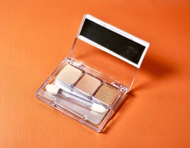 Three neutral brown colored eye shadow make up palette with mirror and brush applicator tool isolated on horizontal orange brown background. clipart