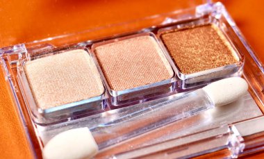 Close up three neutral brown colored eye shadow make up palette with brush applicator isolated on horizontal orange brown background. clipart