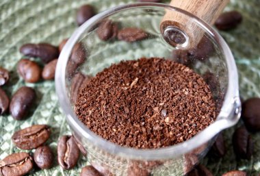 Topraklanmış kahverengi aromatik kahve, organik içecek malzemeleri, küçük şeffaf kap nesnesi, yatay yeşil desenli masa örtüsü, kahve çekirdeği süslemeleri..