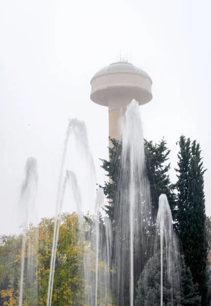 Fountain in the autumn city park with a water tower in the background.