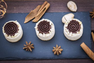 Round white cakes covered with chocolate stripes on a dark stone plate on a wooden table. Decorated with spices and cinnamon sticks