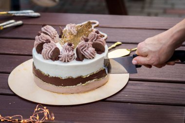 A section of a beautiful cake with layers of different colors and with a decoration of cream and chocolate with golden decor on a stand made of wood with peony flowers in the background