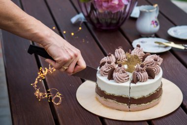 A section of a beautiful cake with layers of different colors and with a decoration of cream and chocolate with golden decor on a stand made of wood with peony flowers in the background
