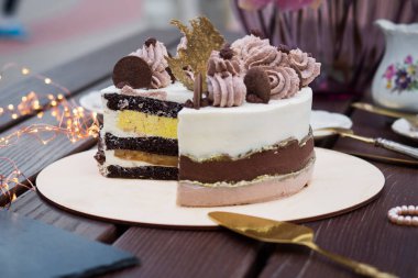 A cut and a piece of a beautiful cake with layers of different colors and with a decoration of cream and chocolate with golden decor on a stand made of wood with peony flowers in the background