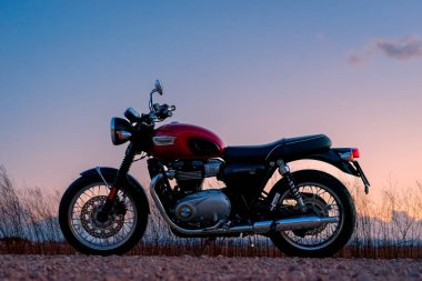 9 Mart 2023 - Albacete, İspanya - Kırmızı Triumph Bonneville T100 motosikleti yol kenarına park edilmiş gün batımı gökyüzü ve bulutlarına karşı