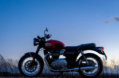 9 Mart 2023 - Albacete, İspanya - Kırmızı Triumph Bonneville T100 motosikleti yol kenarına park edilmiş gün batımı gökyüzü ve bulutlarına karşı