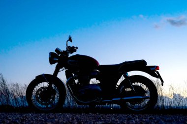 9 Mart 2023 - Albacete, İspanya - Kırmızı Triumph Bonneville T100 motosikleti yol kenarına park edilmiş gün batımı gökyüzü ve bulutlarına karşı