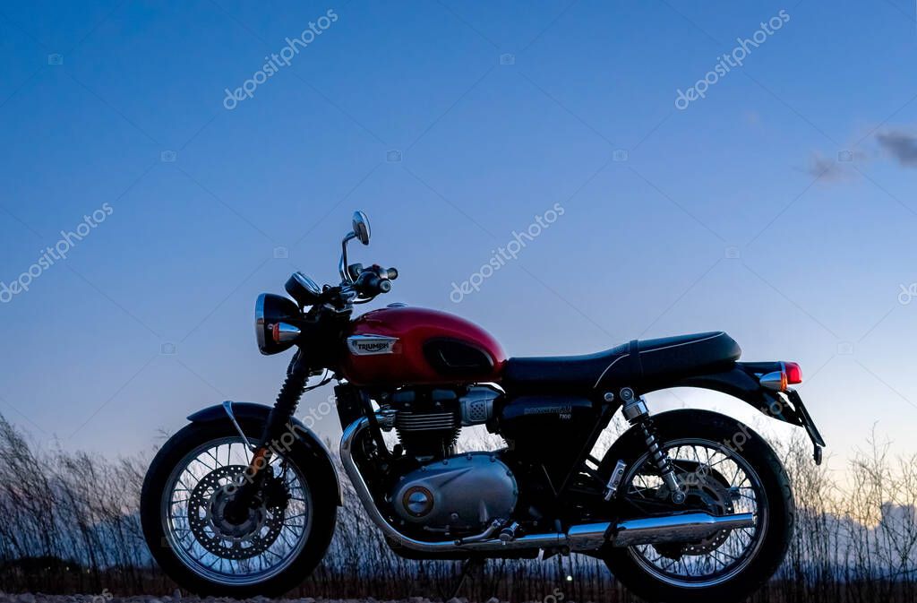 March 9, 2023 - Albacete, Spain - Red Triumph Bonneville T100 motorcycle parked on the side of the road against the backdrop of the sunset sky and clouds