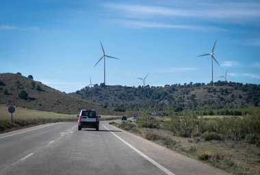 Haziran 2021 - Albacete, İspanya - Yel değirmenlerine tepeden bakan dağ manzaralı bir yolda giden arabalar