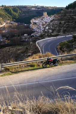 Mart 2023 - Albacete, İspanya - Kırmızı motosikletli Triumph Bonneville T 100 yol kenarında güzel bir İspanyol dağ köyünün arka planında ve gün batımında dolambaçlı bir dağ yolunda