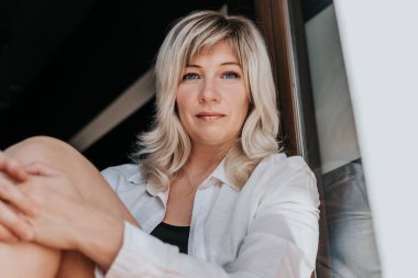 Beyaz bluzlu bir sarışın perdenin arkasından pencereden dışarı bakar.