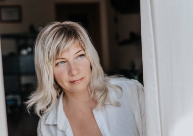 Beyaz bluzlu bir sarışın perdenin arkasından pencereden dışarı bakar.