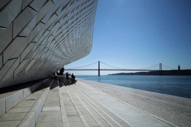 The main and oldest bridge in the city of Lisbon with the modern electricity museum next to the Tagus river. Graphic lines formed by the building and the stairs with people in the shadow. The bridge is from the same architect of the Eiffel Tower.