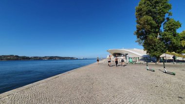 Cityscape in Lisbon, near the Tagus river, with a modern museum, the electricity museum, and a few people running. The sky is blue and the water is dark blue.