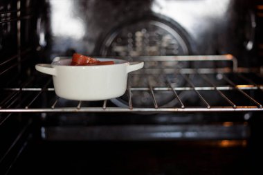 Small white pot inside the oven, with tomato on the inside. The oven is on the grill. 
