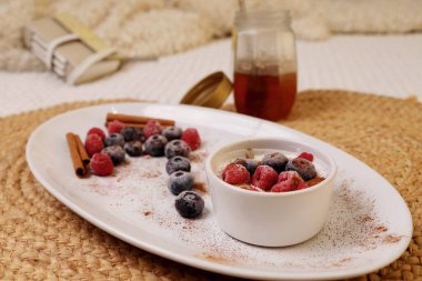 Close up of white bowl with yogurt and red fruits with honey and cinnamon. A white plate with raspberries and blueberries, cinnamon sticks and powder. In the background a jar of honey with the lid off.