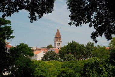 Tarihsel bir saat kulesinin büyüleyici bir görüntüsü, sanatsal çerçevesi ön plandaki verimli yeşillik tarafından, kentsel mimariyi mükemmel bir şekilde harmanlıyor..