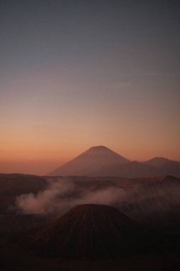 Yükselmiş bakış açısına göre, Bromo 'nun gündoğumu dikey olarak, ılık renkleri gökyüzünü kucaklarken, narin bulutlar aşağıda dinleniyor..