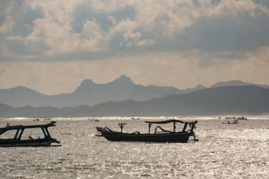 Bu sakin sahnede, Lombok 'un sakin denizinde, heybetli dağlar ve yumuşak bulutlu gökyüzü tarafından çerçevelenmiş bir tekne huzur içinde süzülüyor..