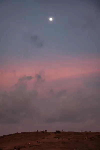 Bukit Merese 'in altından, alacakaranlık tuvaline hayranlıkla bakın. Pembe bir gökyüzü ve yumuşak bir ay doğumu ile süslenmiş. Siluetli insanlar insan varlığı katar.