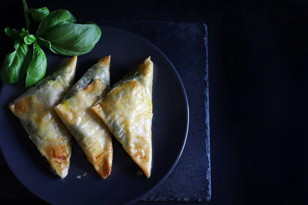 stock image Traditional Greek cuisine. Spanakopita, crispy phyllo pastry triangles with spinach, feta cheese on black background. Directly above. Copy space.