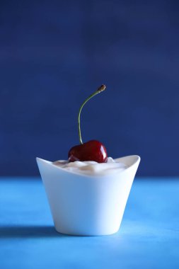 Yaz meyvesi. Mavi arka planda taze kirazlı beyaz bir fincan yoğurt. Boşluğu kopyala, yakın plan.