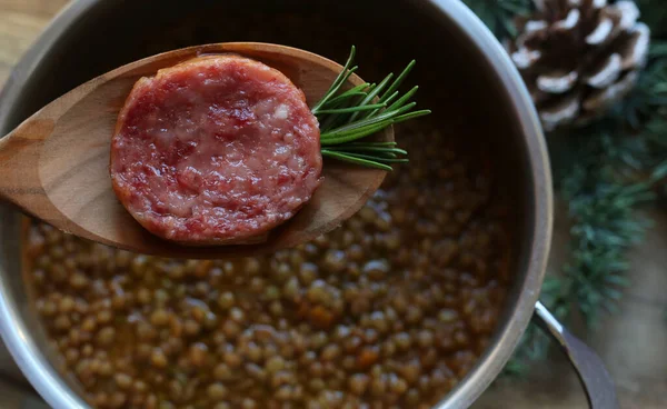 New Year\'s Eve meal Italian style. A slices of cotechino (pork sausage), lentils and rosemary on spoon, blurred background. Overhead view, close-up. Holiday season.
