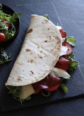 Geleneksel İtalyan piadinası, jambonlu, peynirli, salatalı ve domatesli siyah arka planda. İtalyan yemek sokağı. Genel bakış. Boşluğu kopyala.