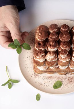 Tiramisu, İtalyan tatlısı. Beyaz arka planda taze nane yapraklarıyla tiramisu süsleyen bir kadın. Genel görünüm.