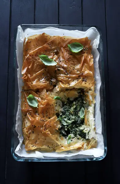 Greek food concept. Spanakopita, delicious greek spinach feta cheese pie on white paper. Dark background. Top view or flat lay.