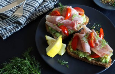 Fırında jambonlu avokado tostu (prosciutto) ve siyah arka planda domates. Sağlıklı gıda konsepti. Genel Görünüm.