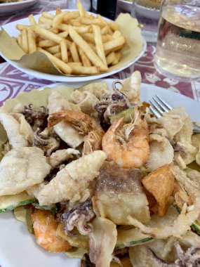 Kızarmış karides, kalamar ve sebze, patates kızartması ve bir İtalyan restoranının arka planında bir bardak beyaz şarap. Tam üstte.