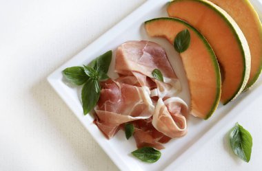 Beyaz arka planda izole edilmiş taze fesleğen yapraklarıyla süslenmiş jambon ve kavun. Yaz yemeği. Tam üstte.