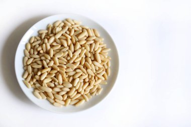 Beyaz arka planda izole edilmiş beyaz bir tabakta çam fıstığı ya da pignoli. Çam kozalaklarının yenilebilir tohumları. Tam üstte. Boşluğu kopyala.