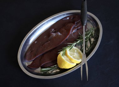 Çiğ ciğer, limon ve siyah arka planda bir tabakta bitkiler. Tam üstte. 