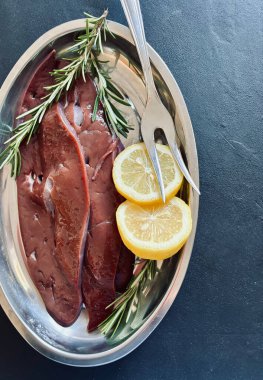 Çiğ ciğer, limon ve gri arka planda bir tabakta bitkiler. Tam üstte. 