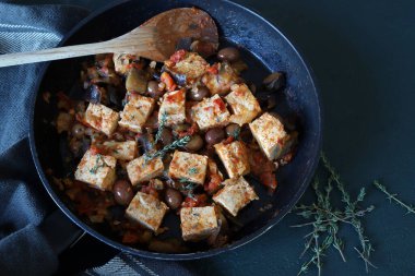 Siyah arka planda domatesli, zeytinli ve otlu tofu kızartması. İtalyan yemeği, putanesca tofu. Vejetaryen ve vejetaryen yemekleri. Sağlıklı yemek..