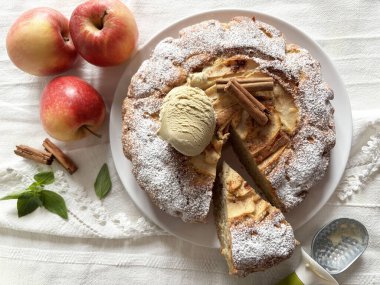Tarçın çubuklarıyla süslenmiş yumuşak ve lezzetli ev yapımı elmalı turta ve bir kepçe dondurma ve arka planda taze elmalar. Tam üstte.