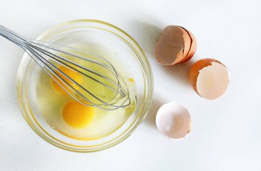 İki çiğ tavuk yumurtası ve cam bir kâse ve beyaz ahşap arka planda izole edilmiş bir el çırpışı. Tam üstte.