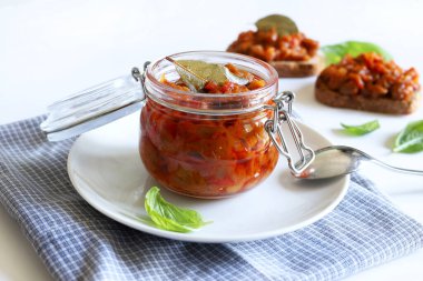 Zacusca - geleneksel Romen yayılımı. Kremalı sebze soslu cam bir kavanoz, patlıcan, soğan, domates ve beyaz arka planda kavrulmuş biber..