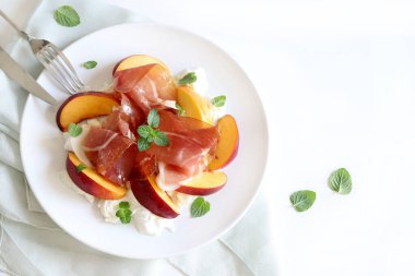 Şeftalili, jambonlu ve burrata peynirli lezzetli bir salata. Mutfak masasında beyaz bir tabakta zeytinyağı ve nane yaprağı ile tatlandırılmış. Tam üstte.