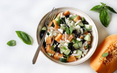 Lezzetli kavun, peynir, zeytin, salatalık aromalı salata ve beyaz bir masada taze fesleğen yaprağı. Hafif bir yaz yemeği. Tam üstte.