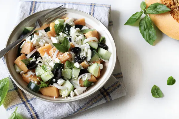 Lezzetli kavun, peynir, zeytin, salatalık aromalı salata ve beyaz bir masada taze fesleğen yaprağı. Hafif bir yaz yemeği. Tam üstte.