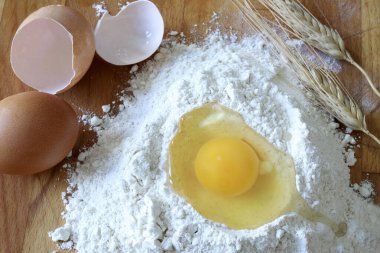 Hamur yapmak için gerekli malzemeler. Kırık yumurtalı beyaz un, bütün bir yumurta, yumurta kabuğu ve ahşap arka planda buğday kulağı. Tam üstte.