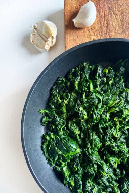 Siyah bir tavada lezzetli ıspanak ve beyaz bir masada sarımsak. Sağlıklı bir yaşam tarzı. Vejetaryen yemeği. Genel Görünüm.