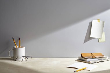 Boş defter, gözlüklü kalem ve duvarın yanındaki masada kırtasiye malzemesi.